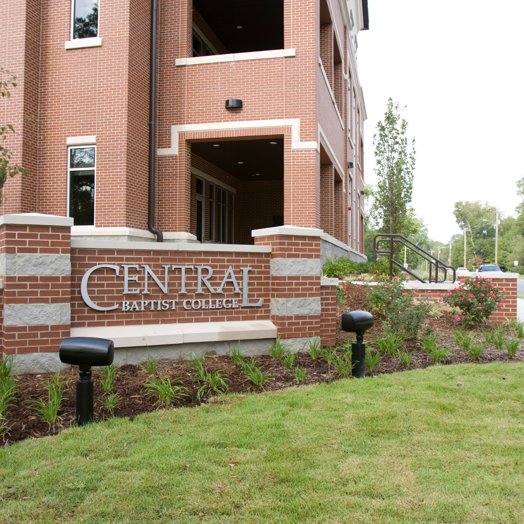 A brick building with “Central Baptist College” displayed on a large sign in front, surrounded by green grass, landscaped plants, and outdoor lights.