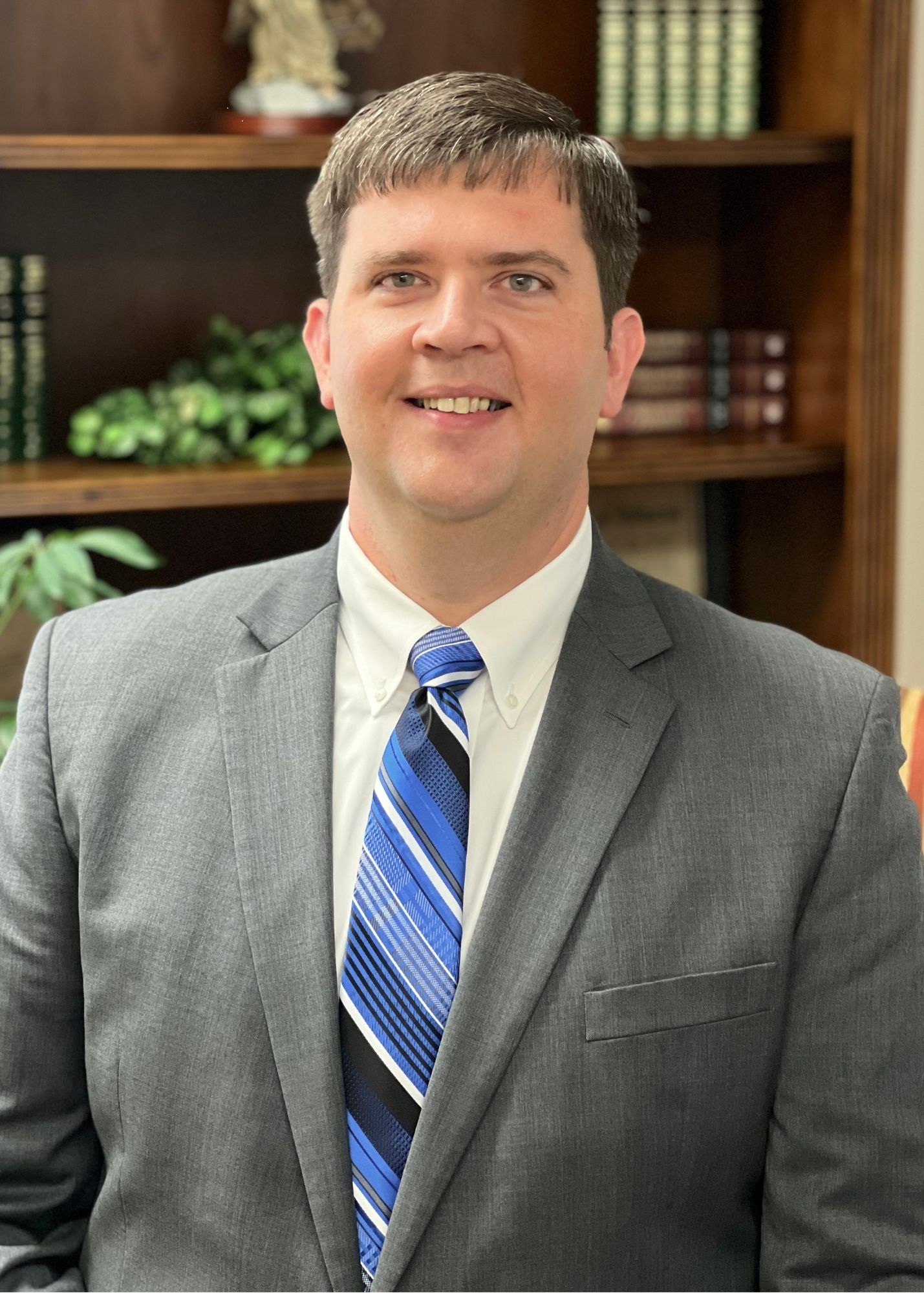 A man in a gray suit, white shirt, and blue striped tie smiles at the camera. He stands in front of a bookshelf with books, a plant, and decorative items in the background.