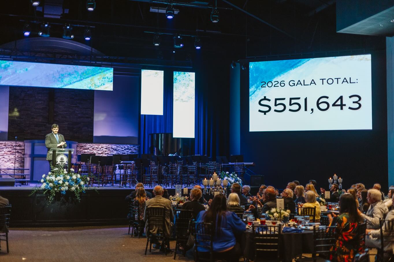 A speaker stands at a podium on stage in front of a seated audience at a gala. A large screen displays 2026 Gala Total: $551,643 in bold text. The setting is dimly lit with blue accent lighting.