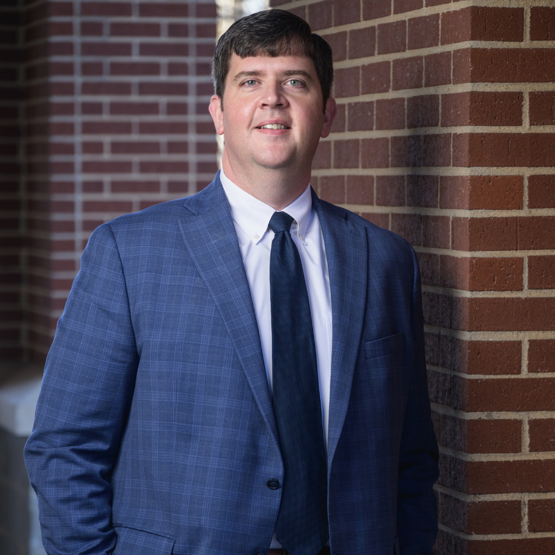 A man in a blue checked suit and navy tie stands by a brick wall, smiling slightly. He has short dark hair and is wearing a white dress shirt. The background features more brickwork and soft lighting.
