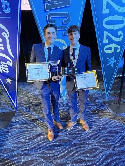 Two young men in suits stand on a patterned carpet, smiling and holding plaques and trophies. Behind them are large blue banners with 2026 and other text, suggesting an award event or competition.