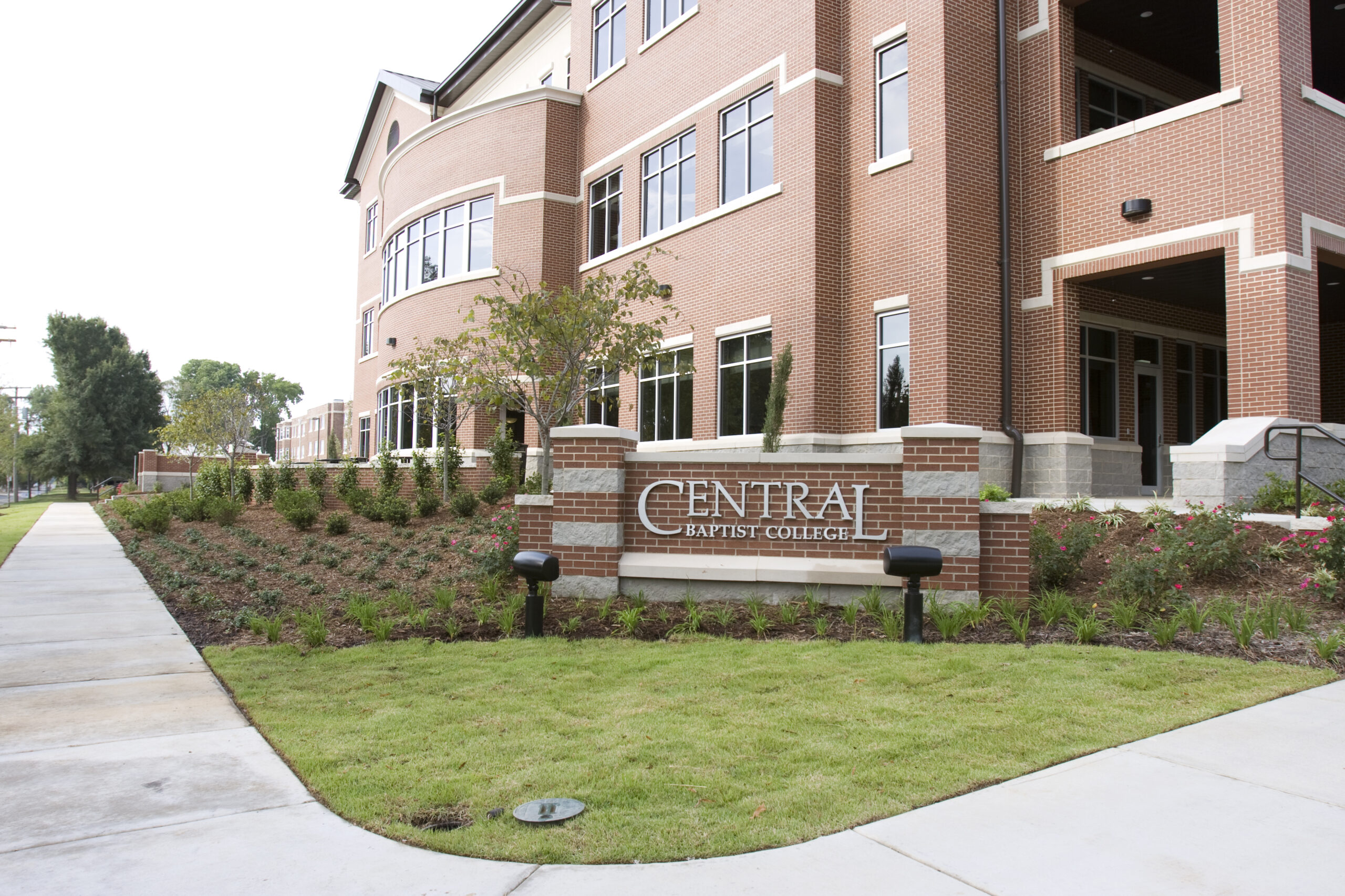 A large brick building with many windows and a sign in front that reads Central Baptist College, surrounded by landscaped greenery and a sidewalk.