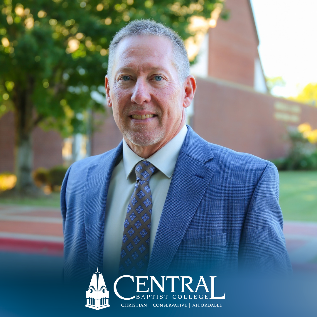 A smiling middle-aged man in a blue suit and patterned tie stands outside in front of the Central Baptist College logo.